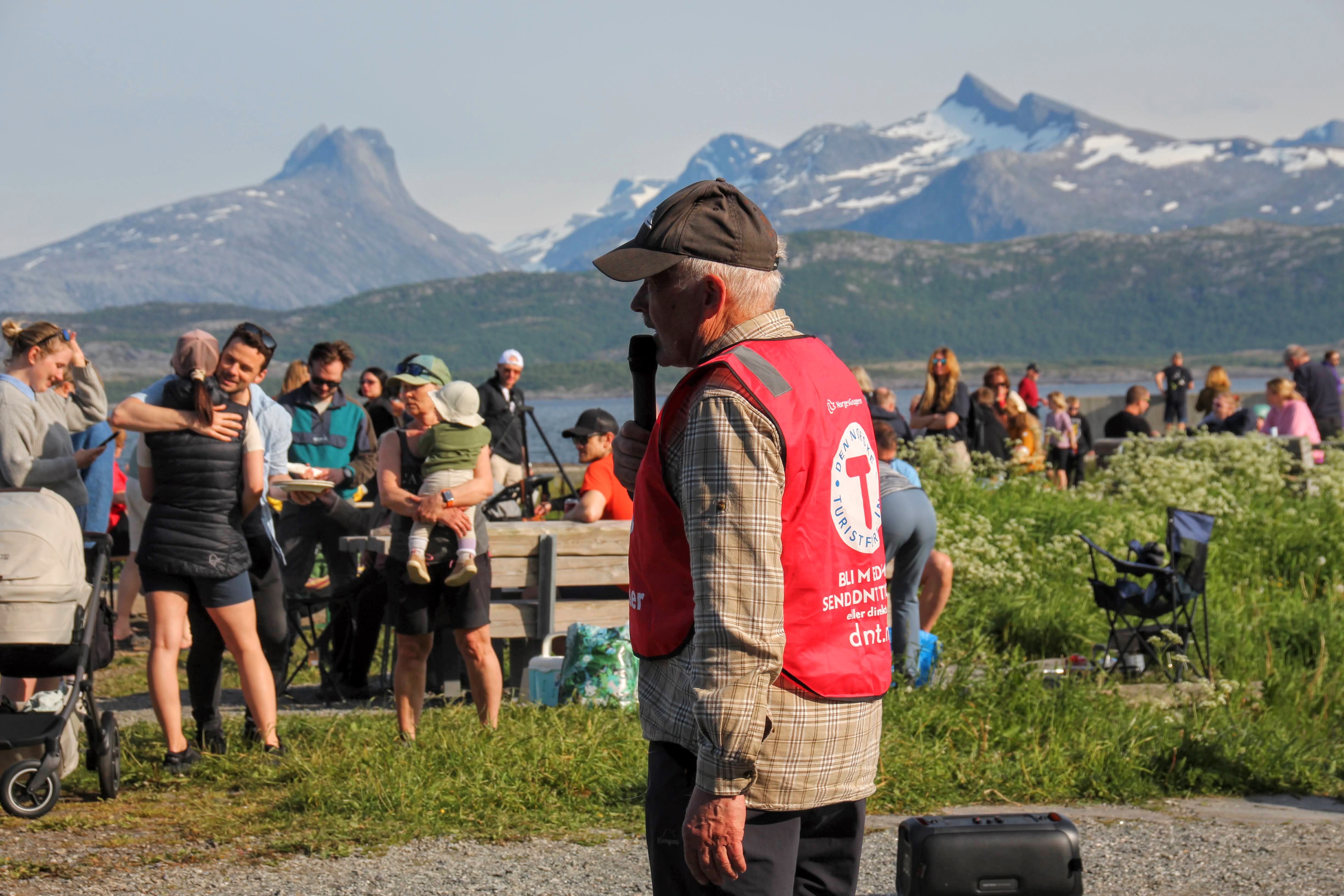 En mann med caps og rød vest snakker i en mikrofon. Det er mange folk i bakgrunnen. Bak er det noen høye fjell.